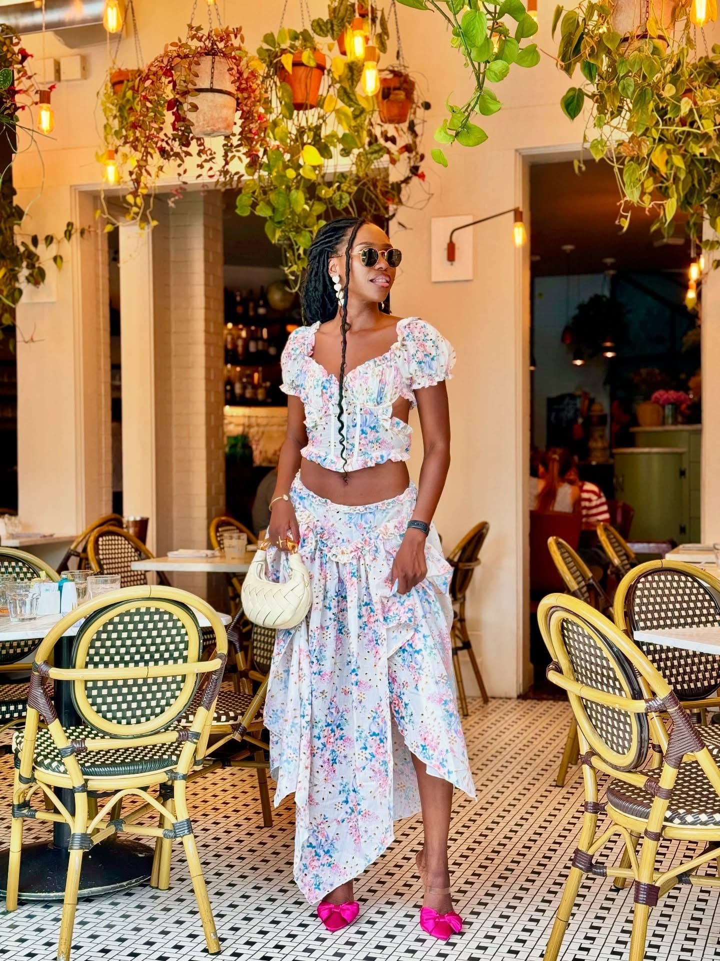 Standing in a plant-filled café in a floral two-piece outfit, sunglasses on, pink bow heels on the tiled floor